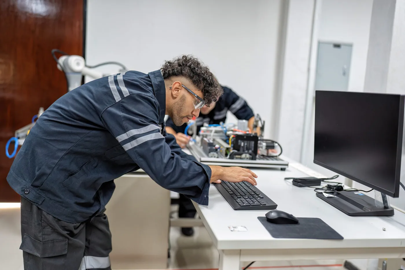 Technicus gebogen over een werkbank met apparatuur en een monitor ernaast